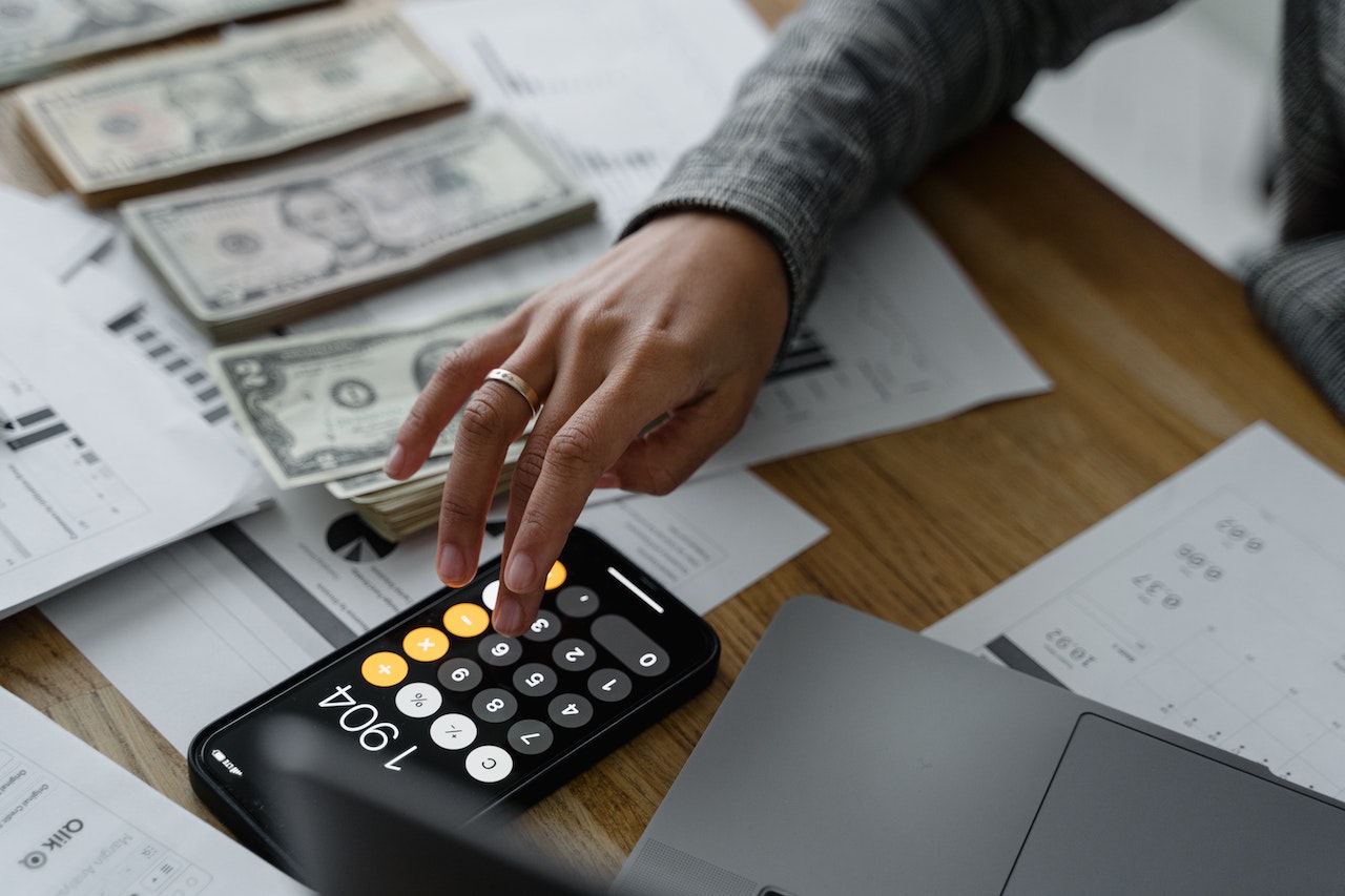 A woman’s hand using a calculator, paperwork, and dollar banknotes lay on the table.